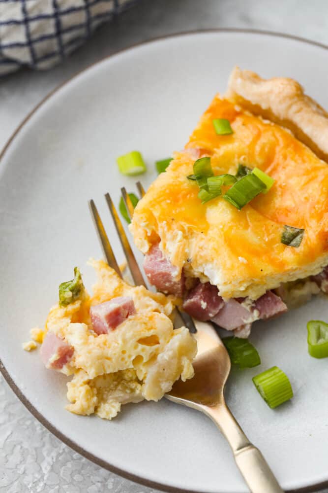 A slice of ham and cheese quiche on a white plate with a gold fork cutting into it. 
