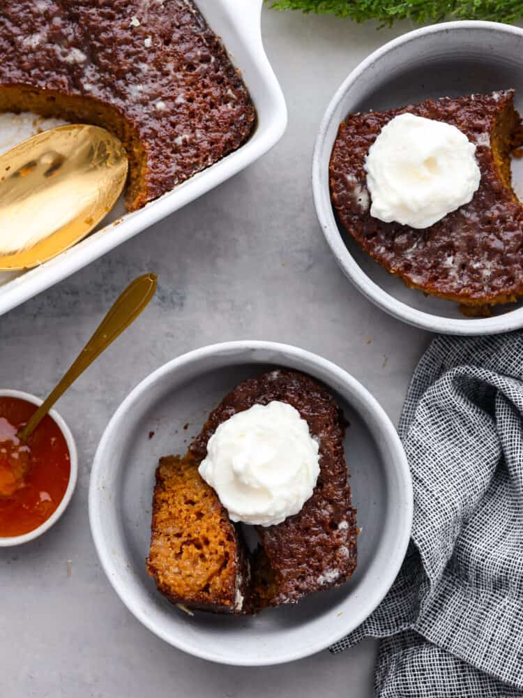 Top-down view of malva pudding served in 2 white bowls with a dollop of whipped cream on top.
