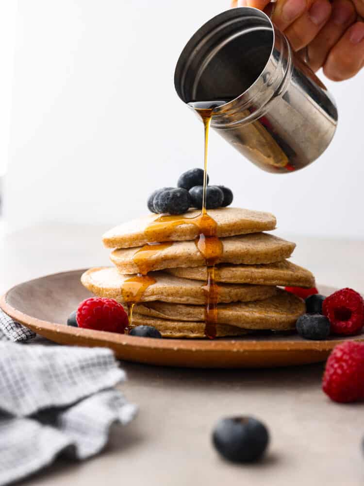 Close view of maple syrup being poured onto the protein pancakes. The pancakes are on a brown plate garnished with fresh blueberries, and raspberries.