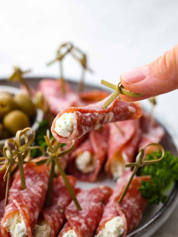 A roll up being held up by a hand over a plate of other salami roll ups.