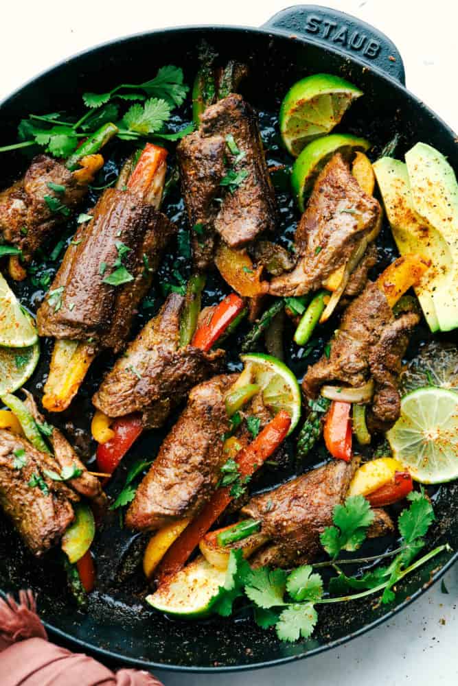 Steak fajita roll ups on the skillet being cooked with lime wedges and cilantro around the skillet for garnish. 