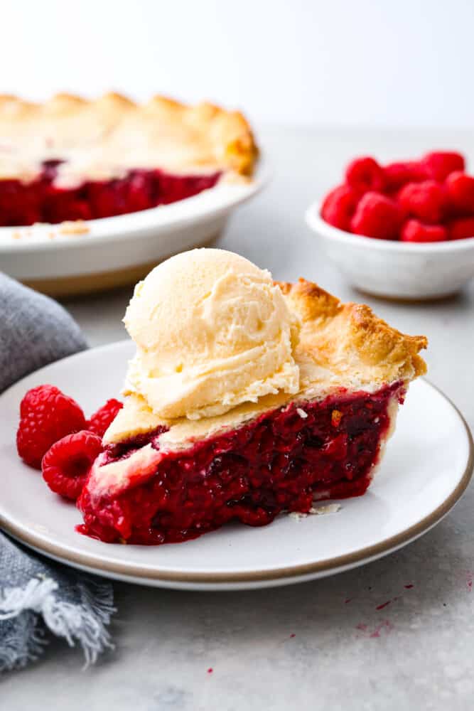 A slice of raspberry pie on a stoneware plate served with a scoop of vanilla ice cream on top.
