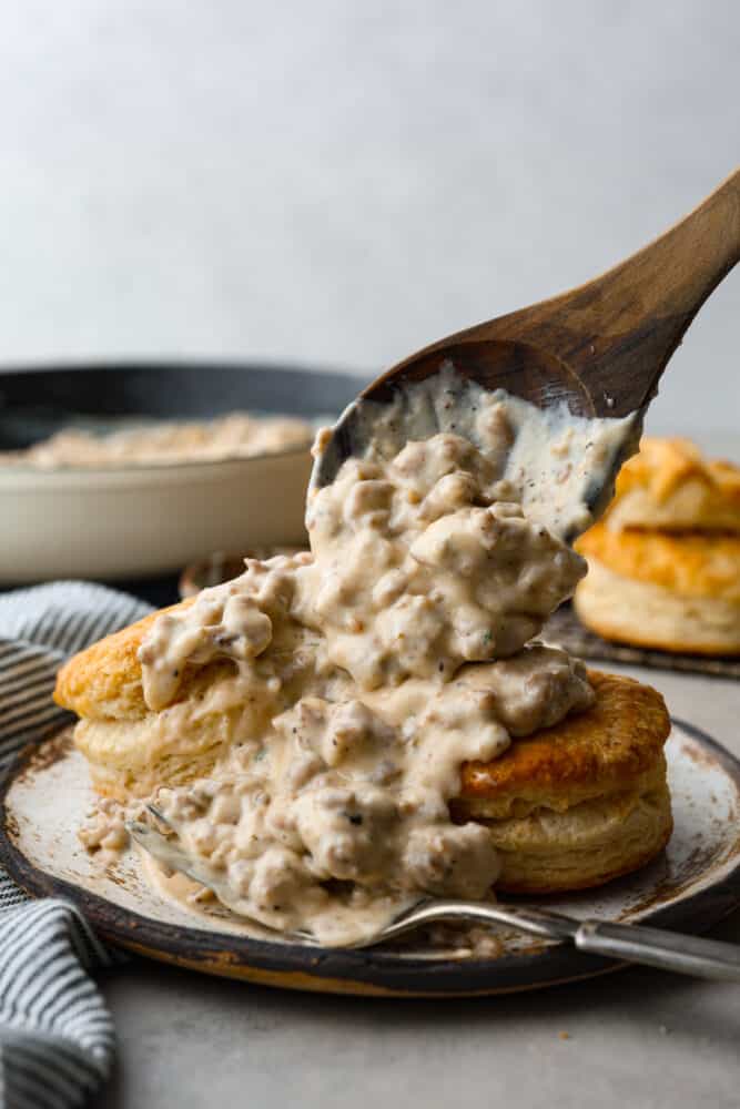 Biscuits and gravy on a plate with a wooden spoon adding more gravy to the biscuits.
