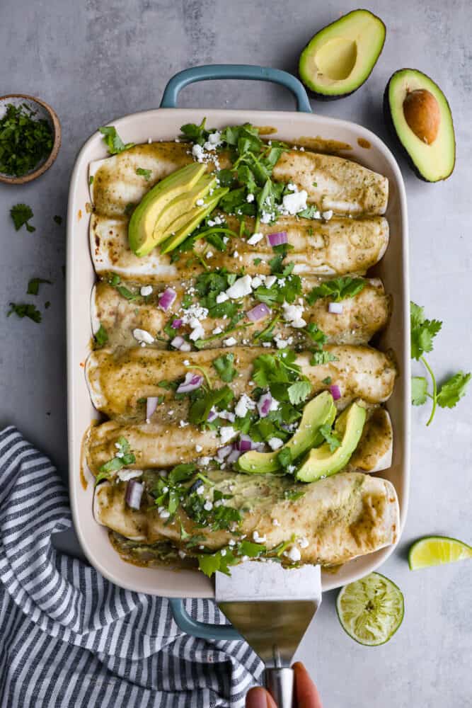 The top few of chicken enchiladas with an avocado cream sauce in a casserole dish with a silver serve about to get an enchilada out. 