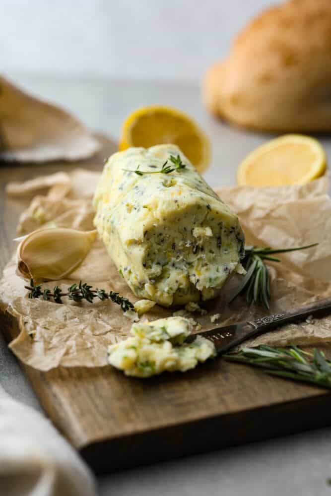 Compound butter on a cutting board with parchment paper, fresh herbs, and a butter knife with butter on it.