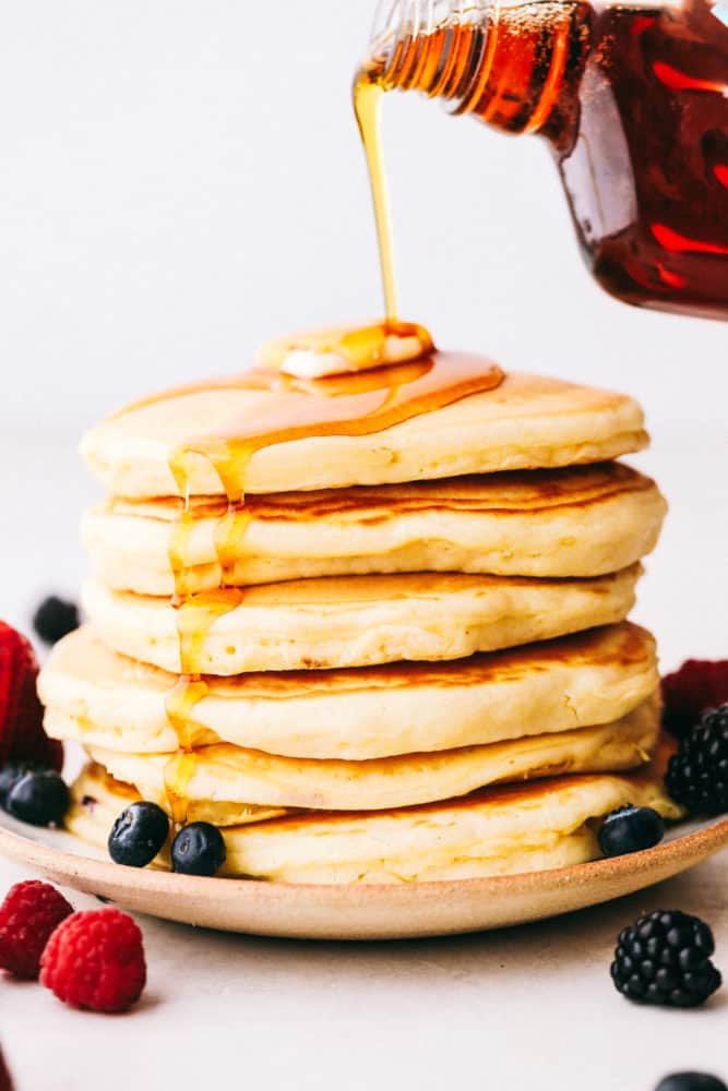 A stack of 6 pancakes with syrup being poured over top of butter and fresh berries. 