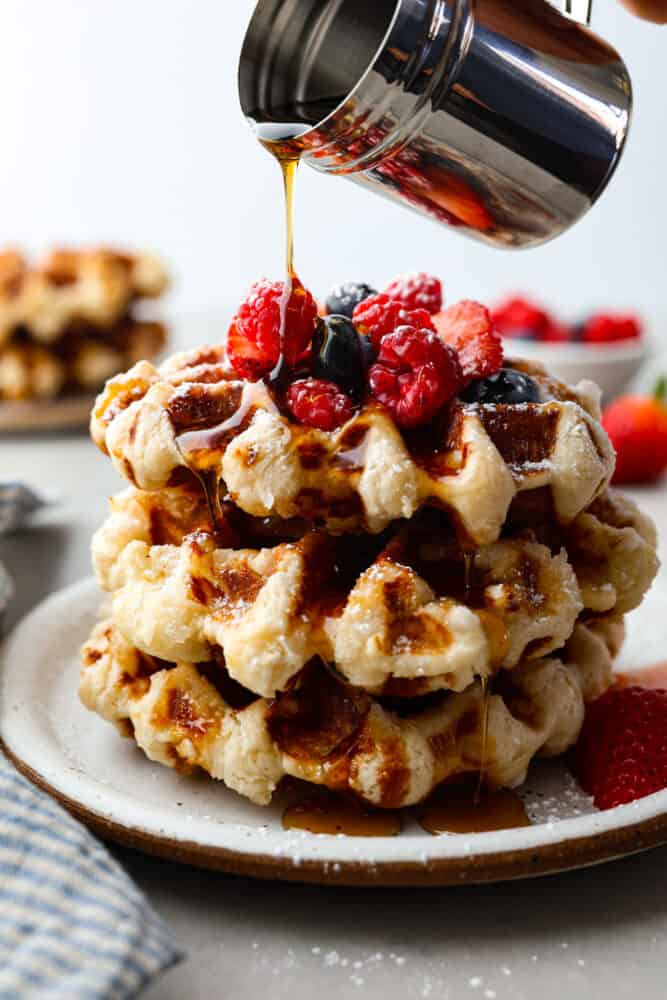 Liege waffles stacked on top of each other, topped with berries, syrup being poured on top.