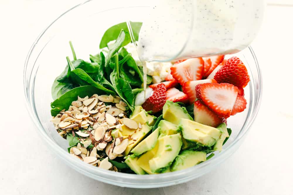 Ingredients to make strawberry avocado spinach salad in a glass bowl with poppyseed dressing being poured over top. 