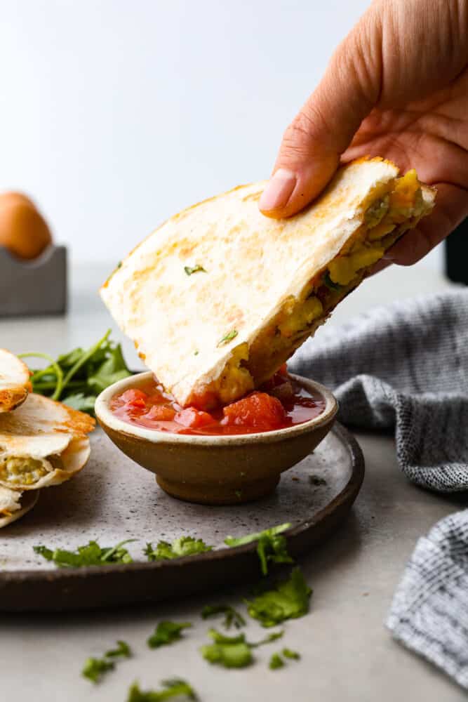 A sliced piece of the breakfast quesadilla is being dipped into a small bowl of salsa.