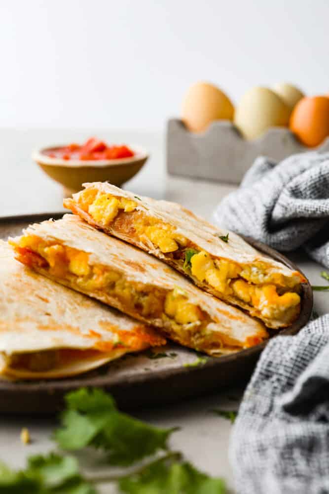 Close up view of breakfast quesadillas stacked on a brown plate. Salsa, fresh eggs, and a blue towel are styled around the plate.