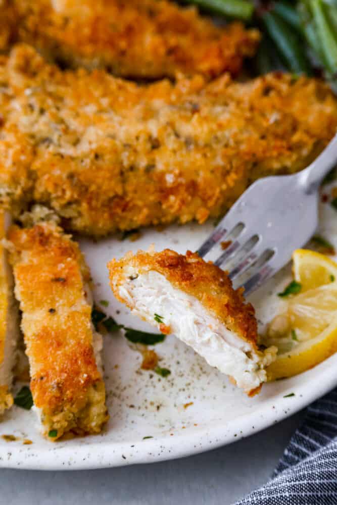 Close up view of chicken Milanese on a white plate. A piece of chicken is on the fork. Green beans and lemon slices are next to the chicken.