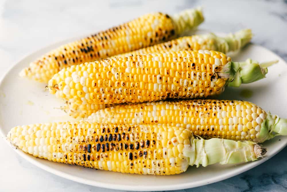 This corn gets grilled and slathered with an amazing mayonnaise blend and topped with parmesan cheese and chili powder. This is one of the best corn recipes you will eat! Grilled corn stacked on a white plate.