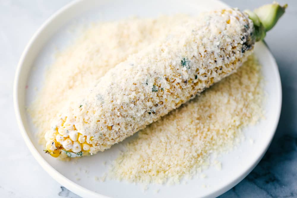 Grilled corn coated with mayonnaise and being rolled in Parmesan cheese.