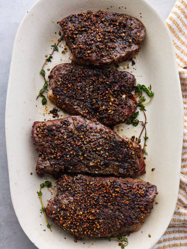 Searching for an impressive, hearty dinner? Look no further than this steak au poivre! This delectable French dish consists of peppercorn-crusted beef tenderloins coated in a garlic herb cream sauce. It’s so flavorful, so delicious, and perfect for any occasion! Top-down view of 4 steaks on a white serving dish.
