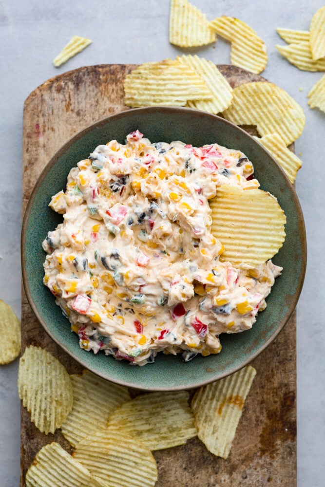A bowl of creamy ranch dip on a wooden cutting bread with potato chips scattered around. 