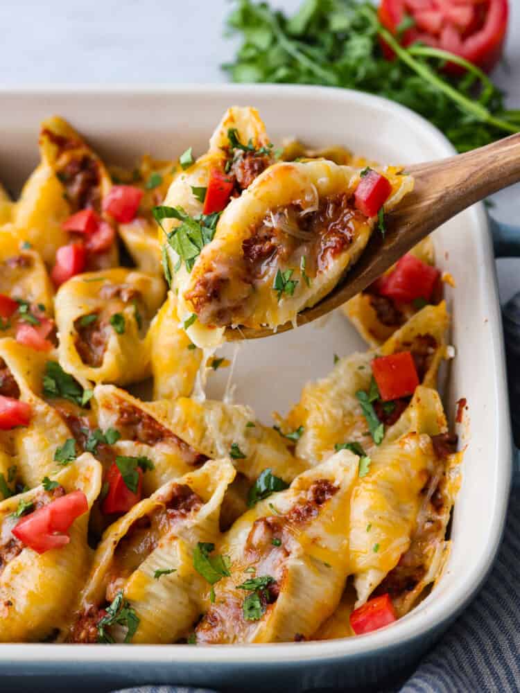 Close view of a wooden spoon lifting up a taco stuffed shell from the baking dish. Cilantro and tomatoes are garnished on top and on the side of the baking dish.