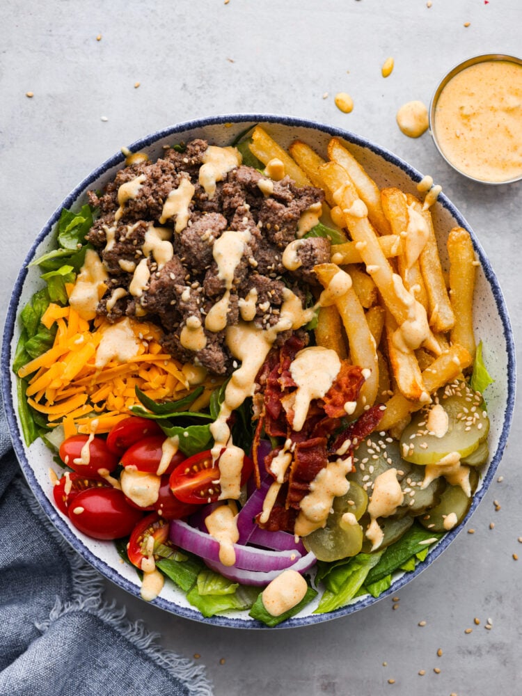 Top-down view of a burger bowl made with beef, bacon, veggies, and shredded cheese, all topped with a creamy sauce.