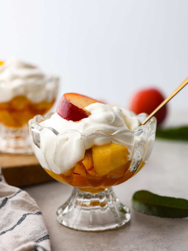 Close view of peaches and cream in a glass dish with a gold spoon. A striped towel and peach leaves are next to the dish.