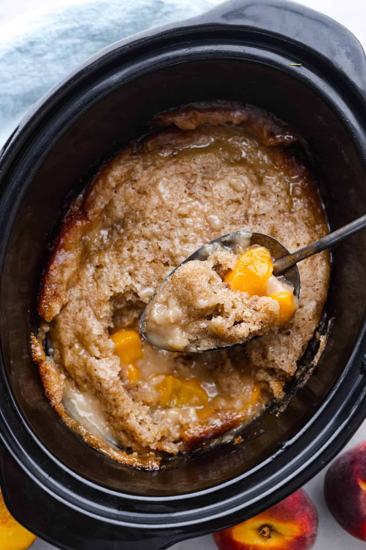 This Slow Cooker Peach Cobbler is an easy dessert, perfect for those fresh summer peaches! It has a sweet biscuit topping and deliciously sweet peach filling underneath that is perfect when served with a scoop of vanilla ice cream. Peach cobbler in a slow cooker being scooped out with a spoon.