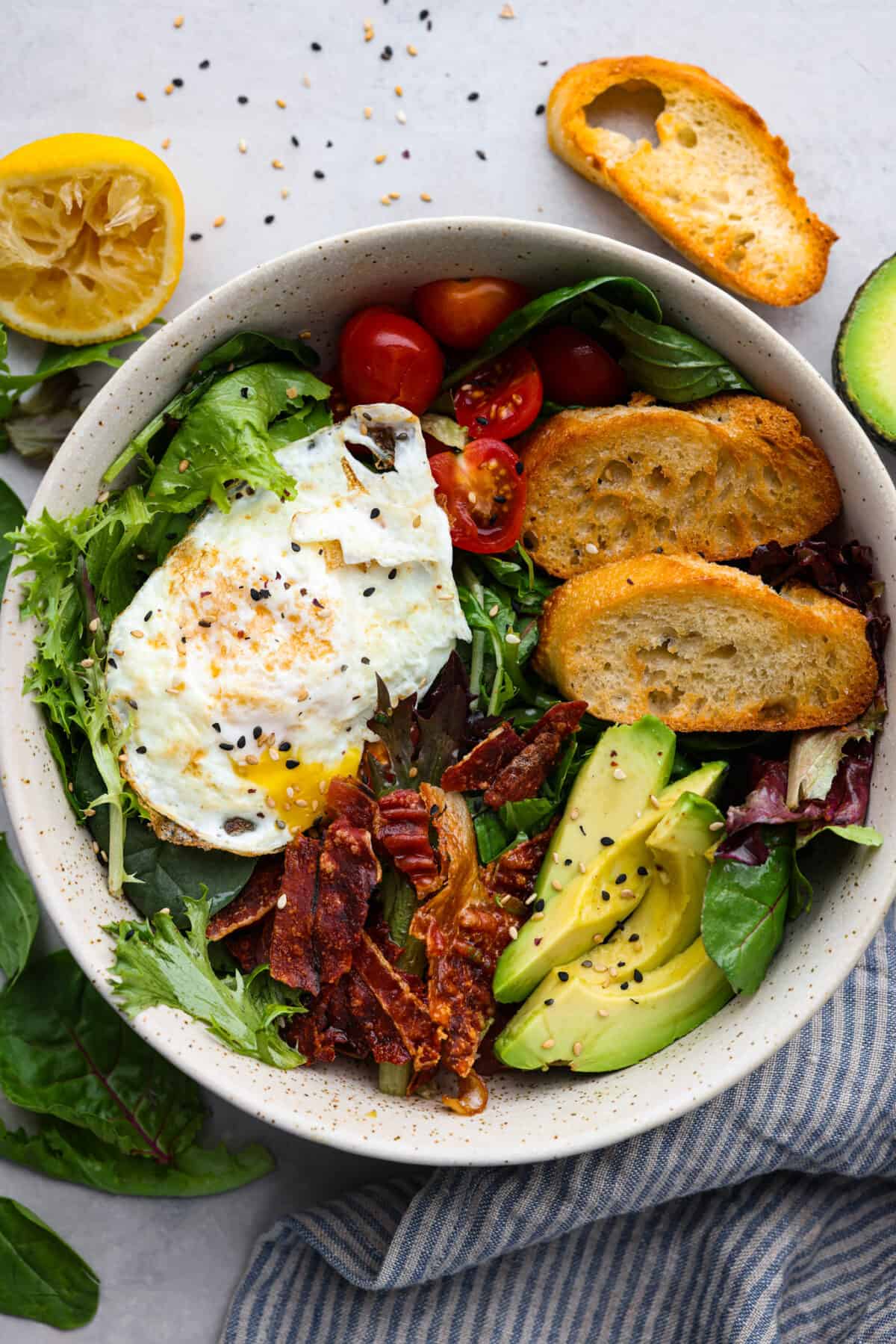 Top-down view of a salad topped with a fried egg, bacon, tomatoes, crostini, and sliced avocado.
