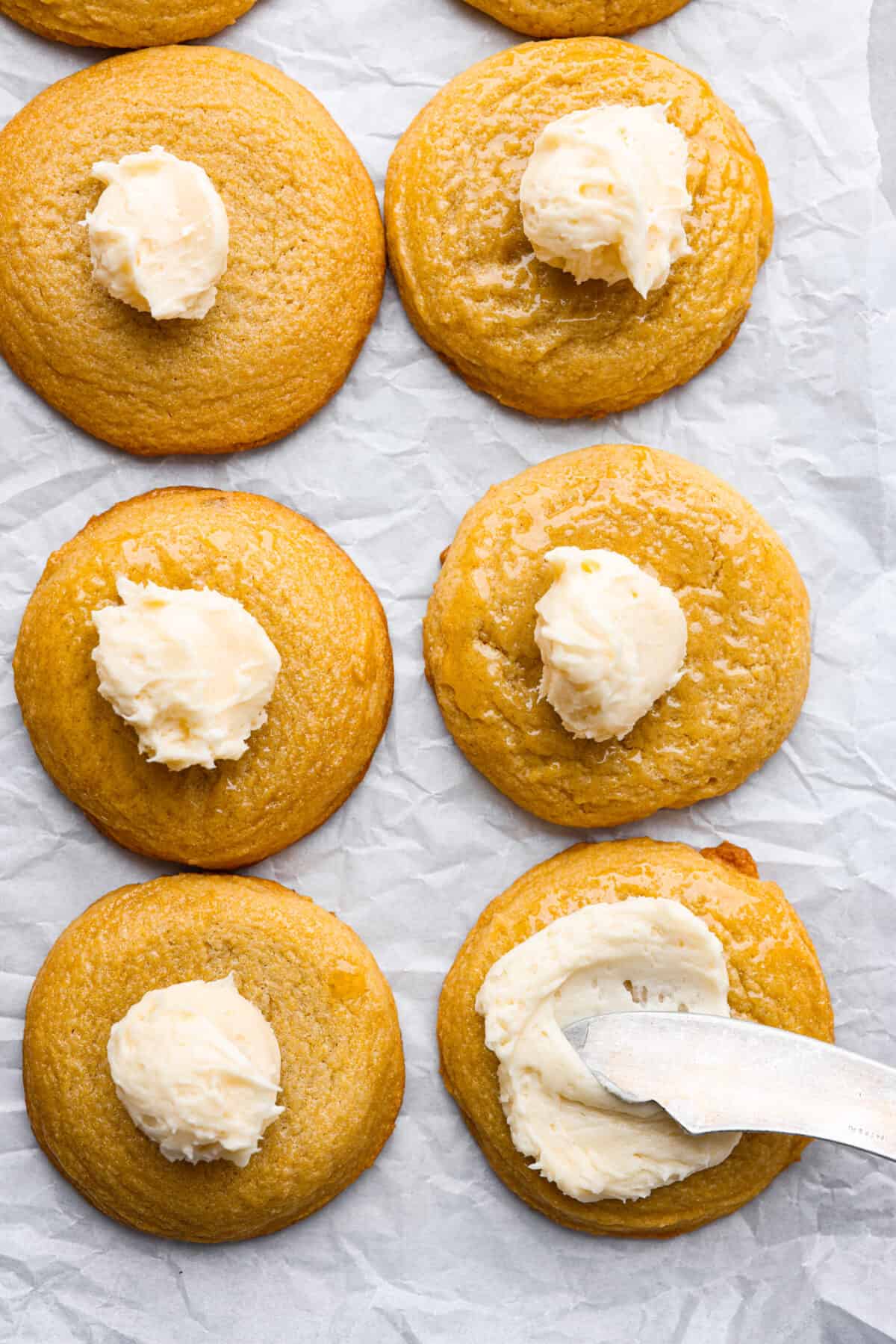 Top view of cornbread cookies with a dollop of frosting on top of each cookie. A knife is spreading the frosting on one of the cookies.