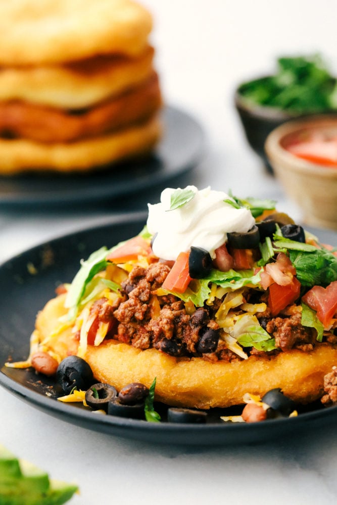 Navajo taco on a black plate with beans, ground beef, tomatoes, lettuce, cheese and sour cream.