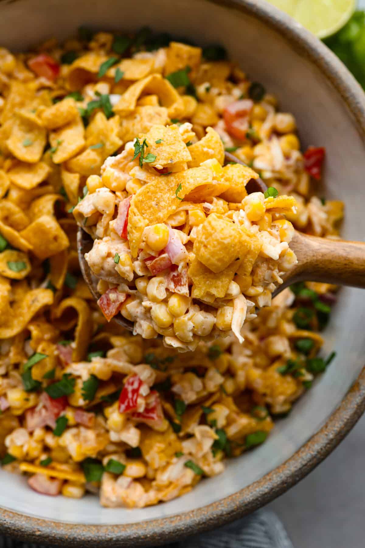 Close view of frito corn salad in a bowl. A wood spoon is lifting up a scoop of salad.