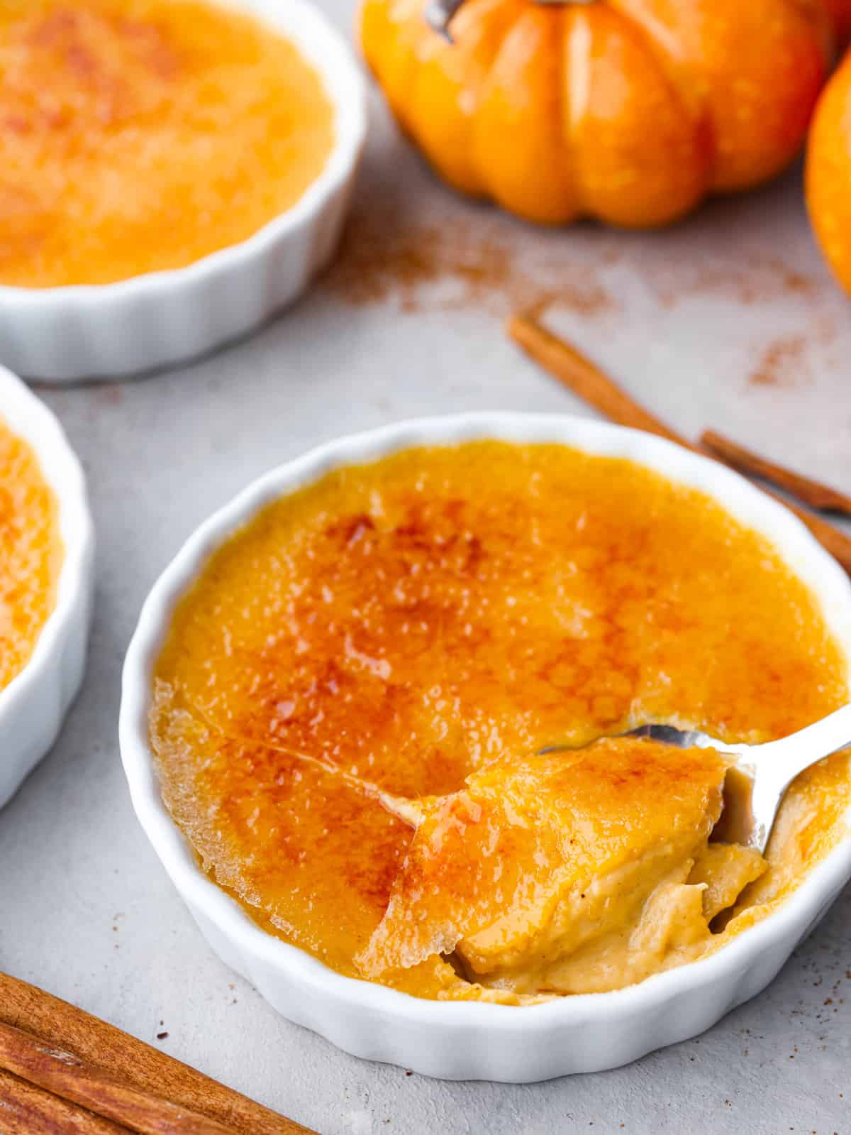 Close side view of a spoon in a ramekin of pumpkin crème brûlée.The caramelized top is cracked into with the spoon. A pumpkin and cinnamon sticks are next to the ramekins.