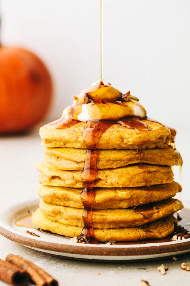 Side view of a stack of pumpkin spice pancakes topped with whipped cream and syrup being drizzled on top.