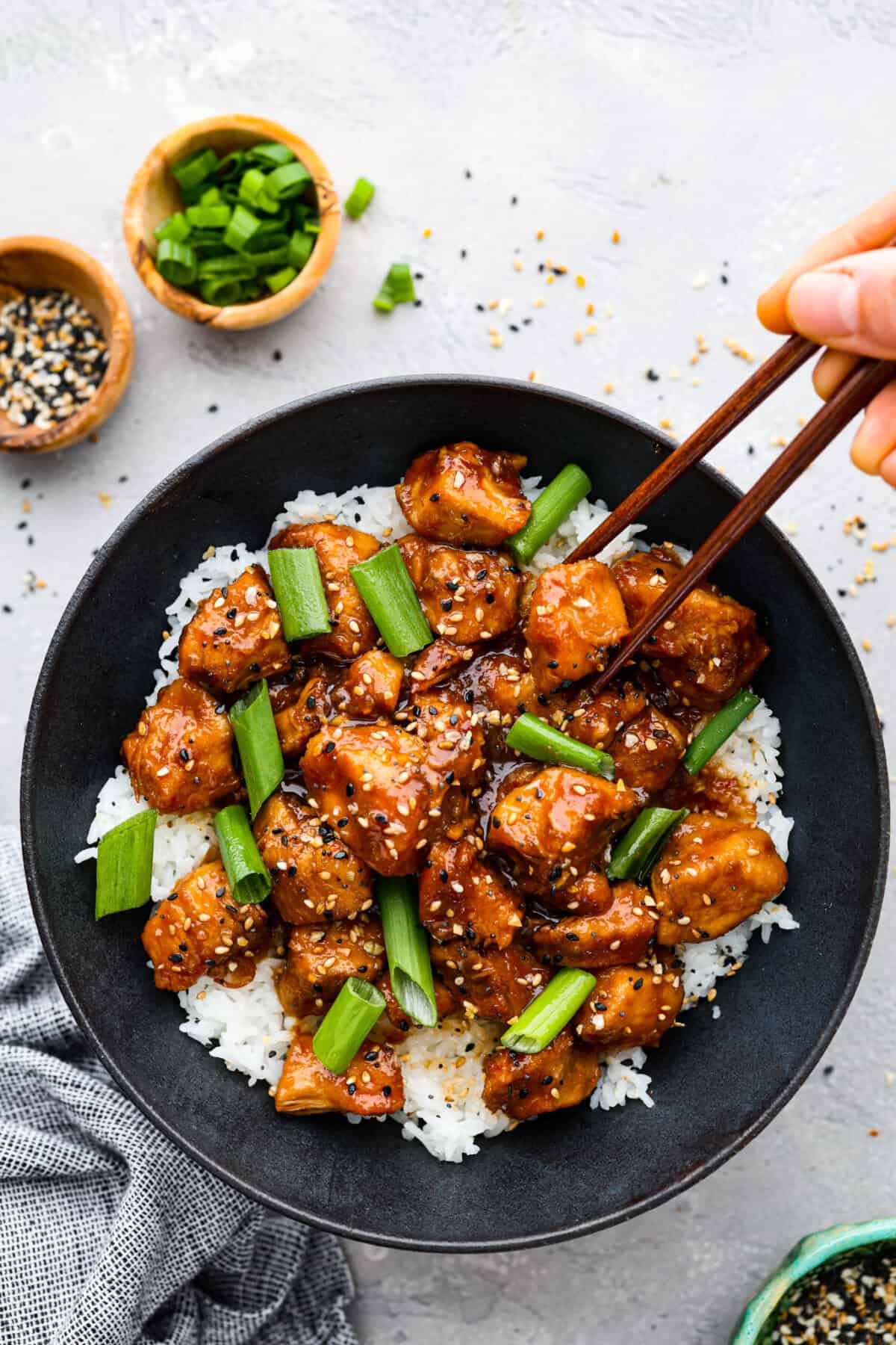 Top view of slow cooker General Tso's chicken in a black bowl served over rice. Garnished with sesame seeds and green onions. Chopsticks are picking up a piece of chicken.