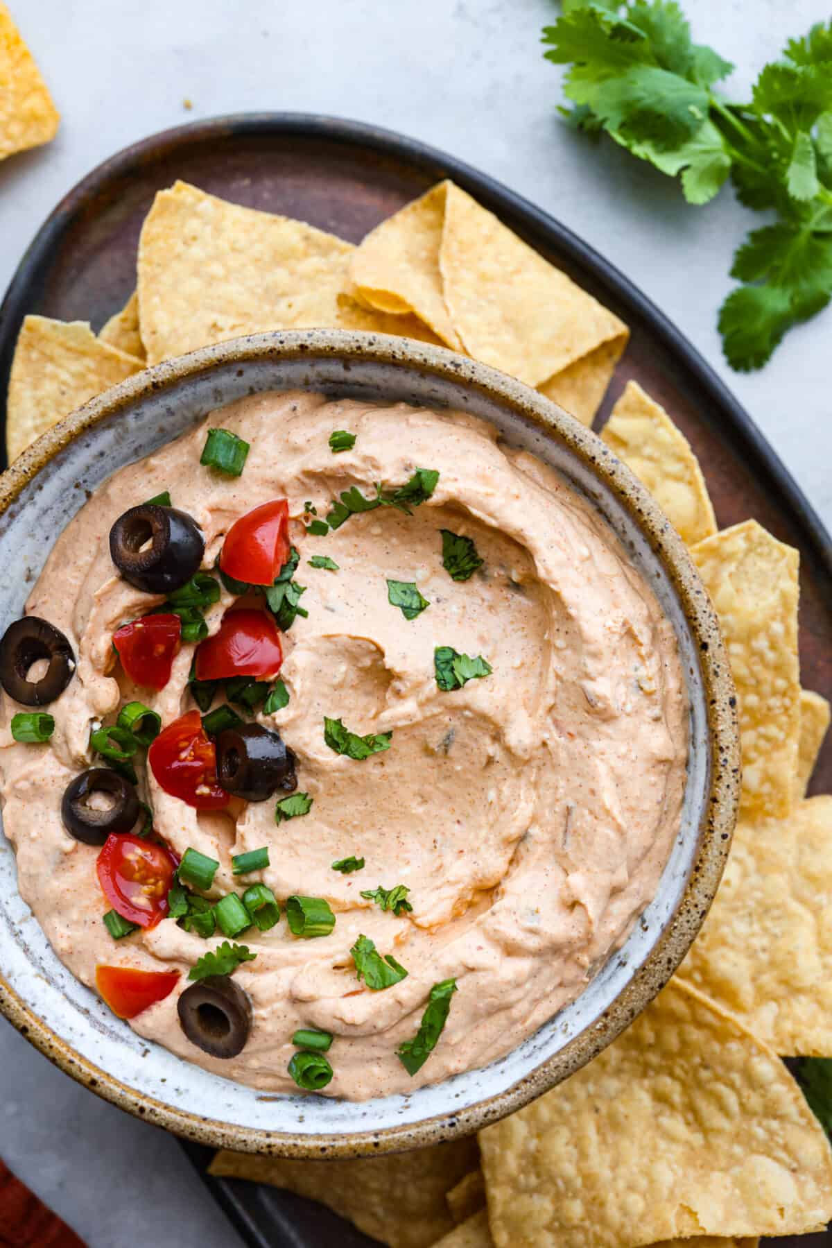 Hero image of taco dip garnished with tomatoes, olives, and cilantro in a stoneware bowl. It is surrounded by tortilla chips.