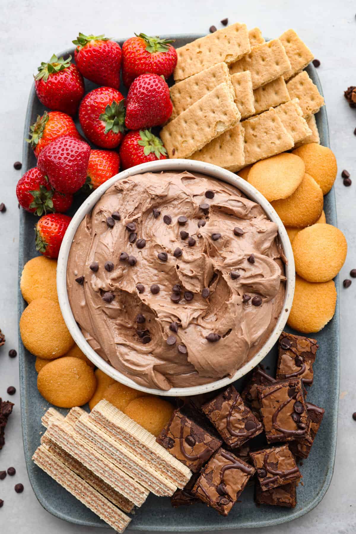 Top-down view of brownie batter dip in a white bowl. It is surrounded by a variety of cookies, cut brownies, wafers, and fruit for dipping. Everything is served on a blue-gray stoneware serving dish.