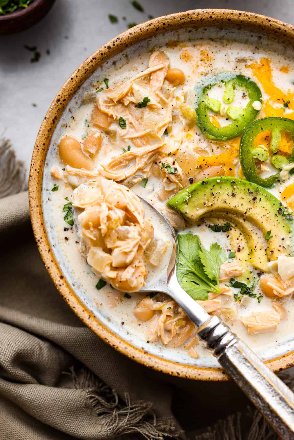 A close up of a bowl of white chicken chili with a spoon and garnished with some jalapeños, avocado slices and some cilantro. 