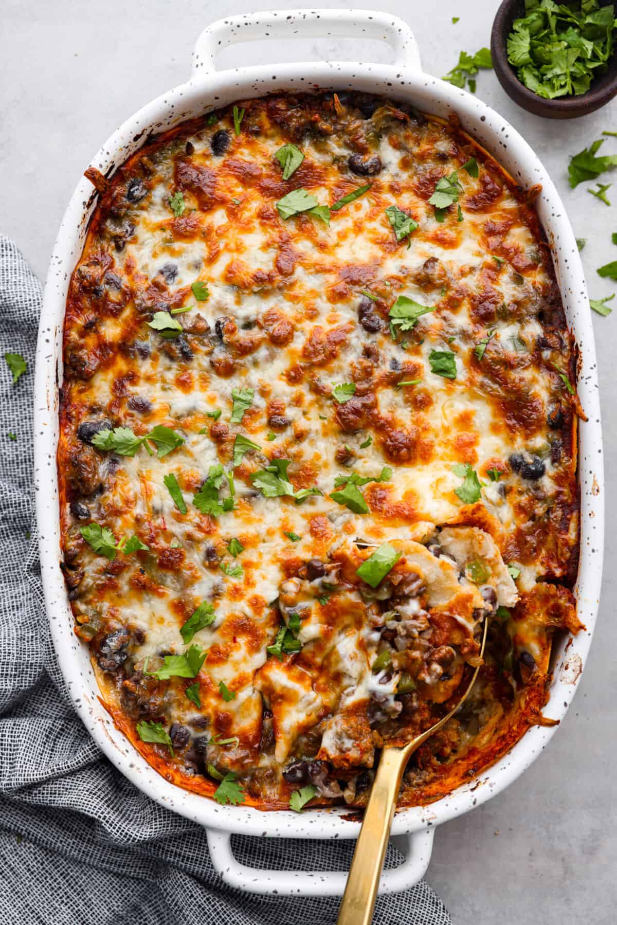 Enchilada Pie is layers of seasoned ground beef, veggies, beans, cheese, enchilada sauce, and corn tortillas; all baked together until perfectly cheesy and golden. This casserole recipe is an easy twist on enchiladas without the hassle of rolling! Top-down view of a baked enchilada pie. There is a golden metal spoon scooping into it.