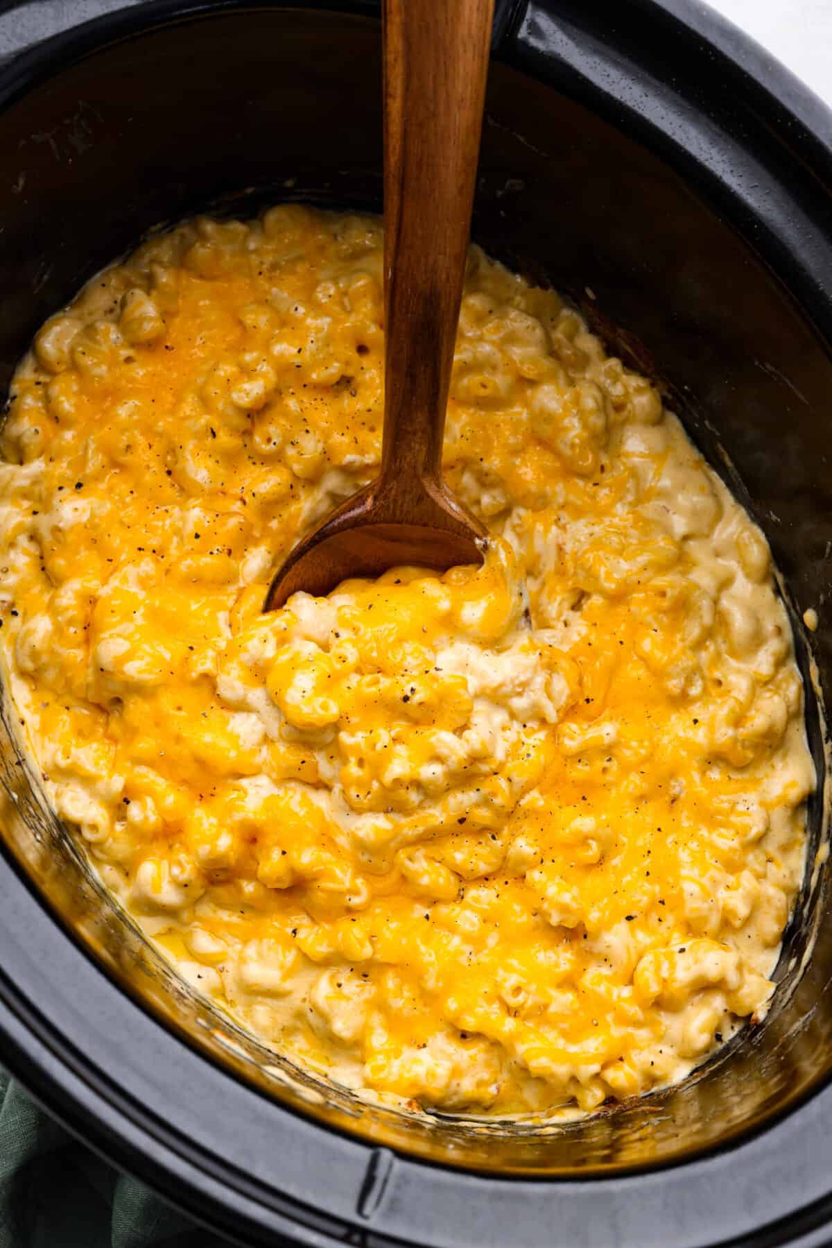 Macaroni and cheese in the bottom of a slow cooker, being mixed with a wooden spoon.