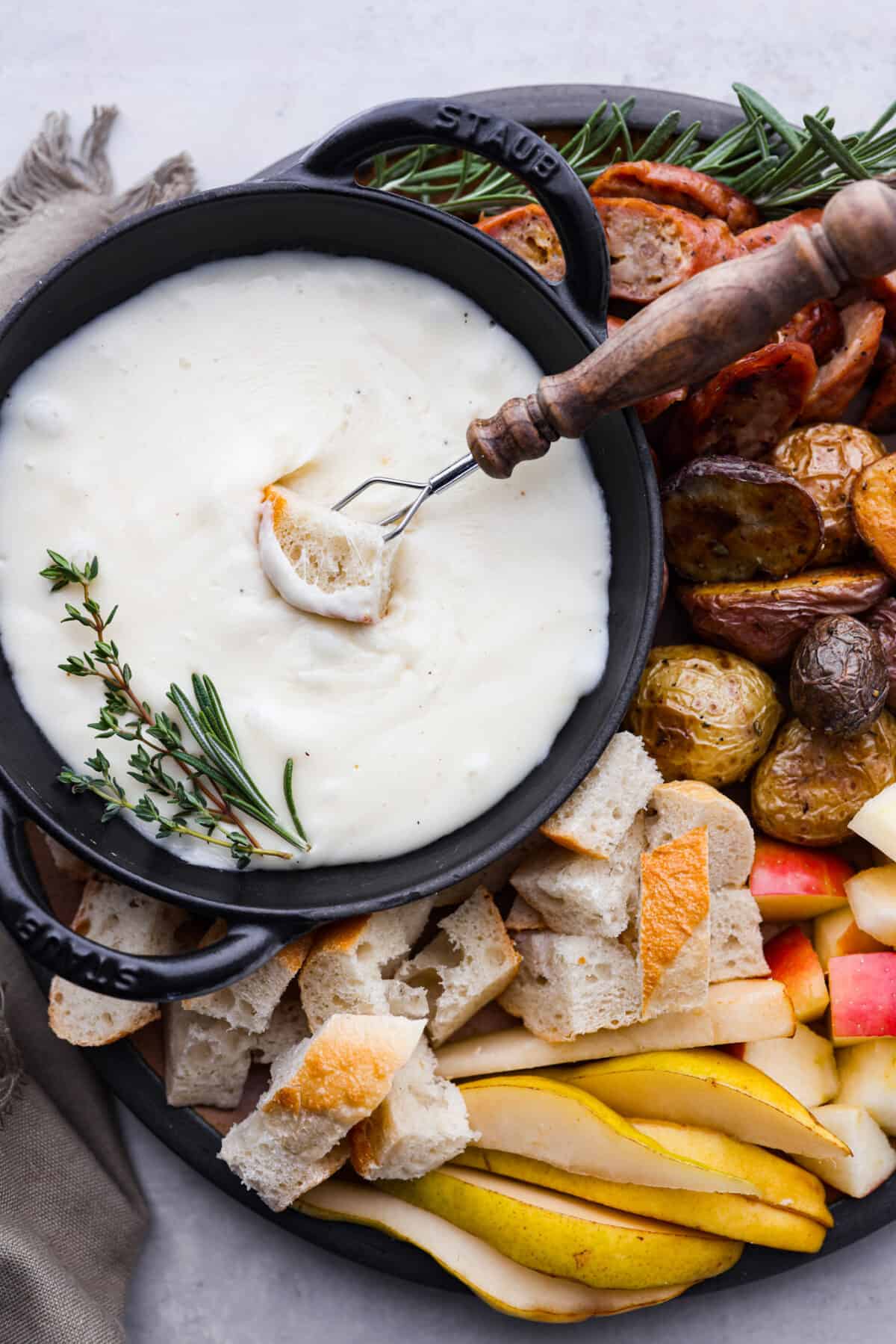 Top-down view of cheese fondue surrounded by bread, potatoes, fruit, and fresh herbs.