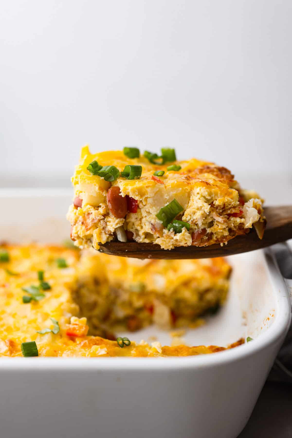 A slice of breakfast casserole being lifted out of the pan with a wooden spoon.