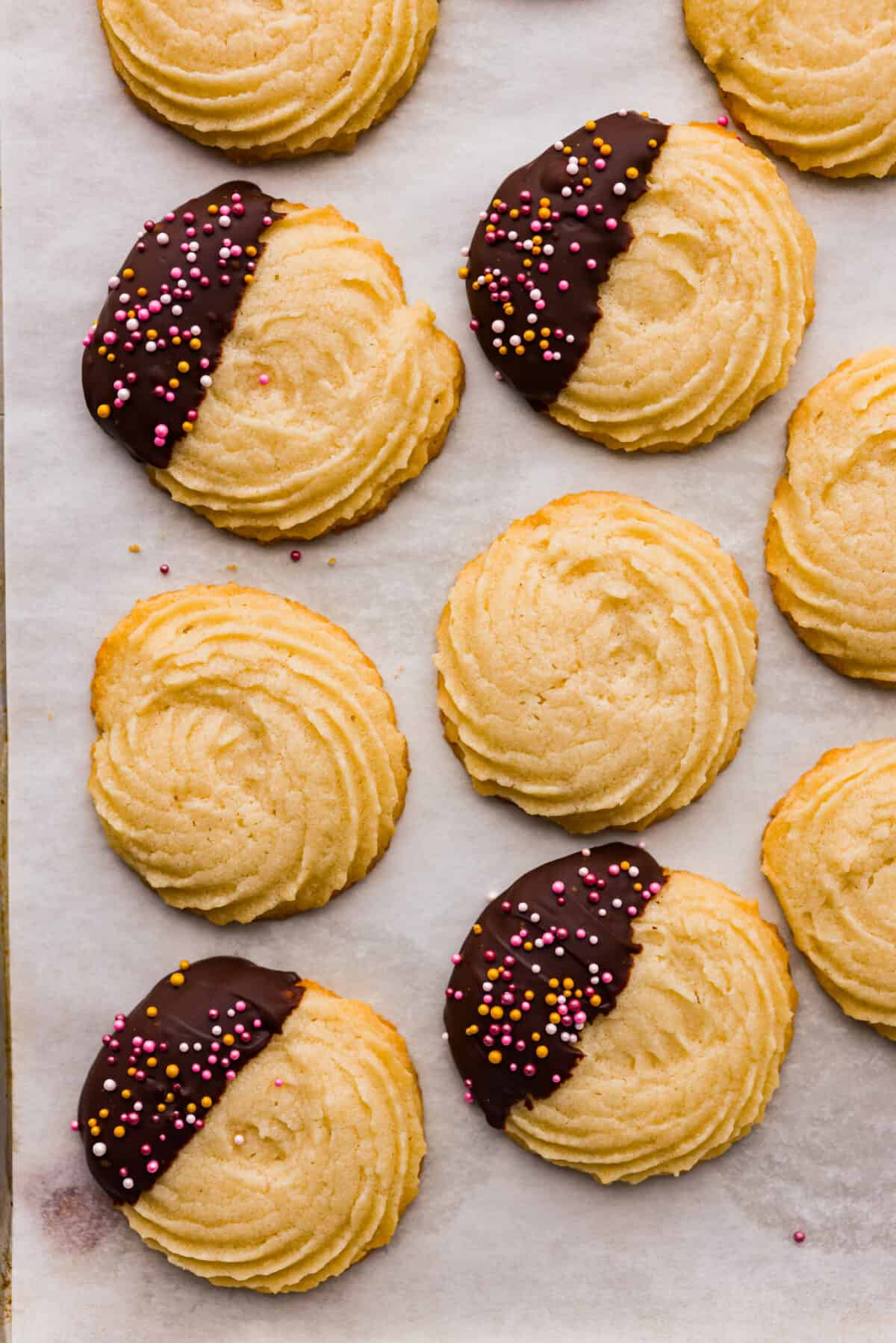 Top close view of butter cookies on a baking sheet pan. Some are plain, some are dipped with sprinkles. 