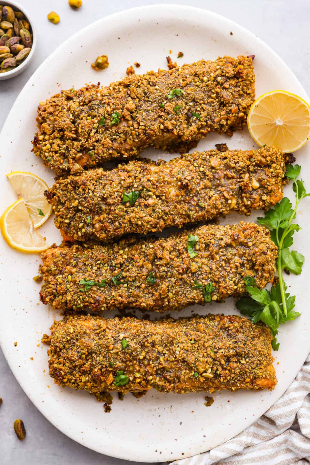 4 pistachio crusted salmon filets on a white plate, garnished with lemon.