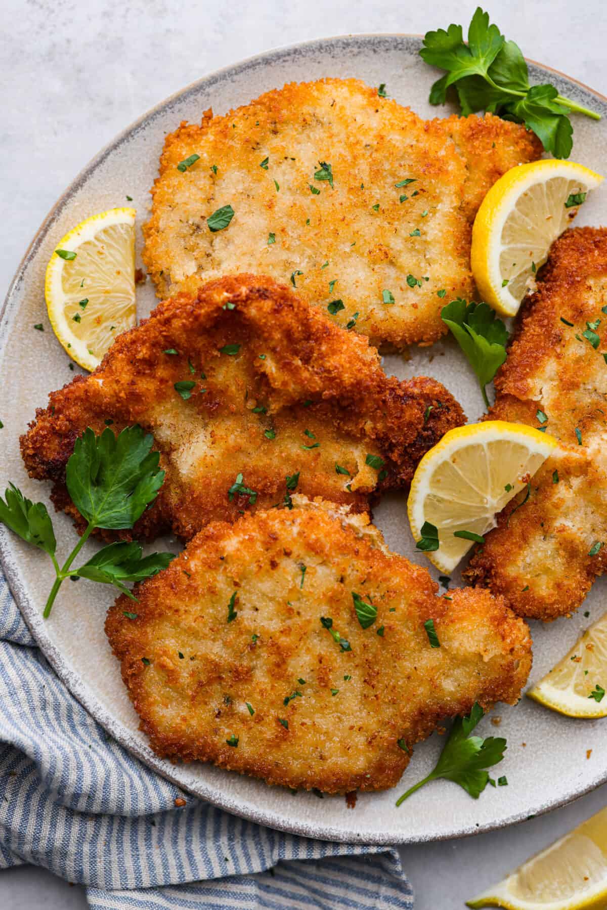 4 breaded pork cutlets on a white plate served with lemon wedges and fresh herbs.