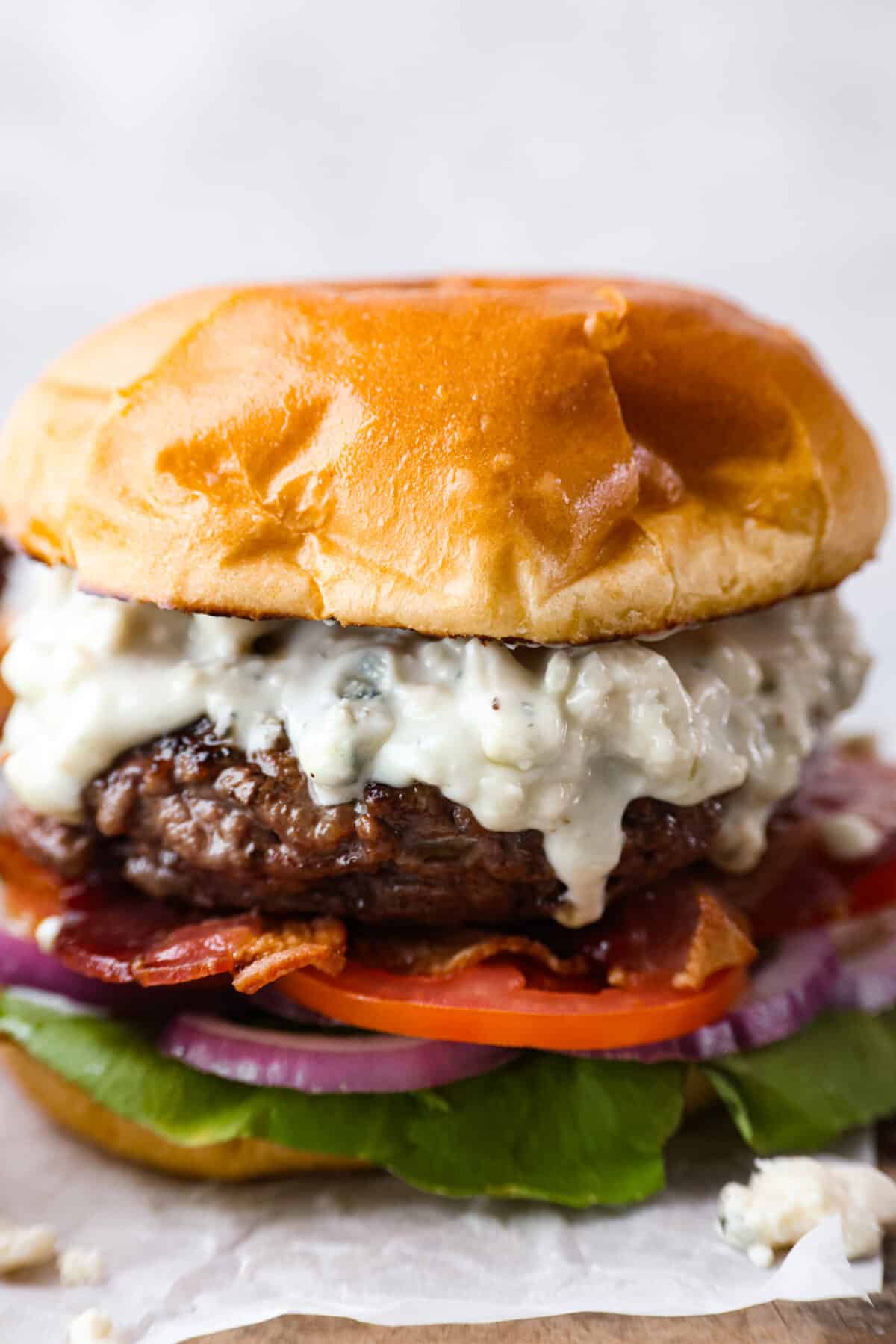 Close up side view of burger dripping with blue cheese dressing, toasted bun, lettuce, tomato, bacon and red onion.