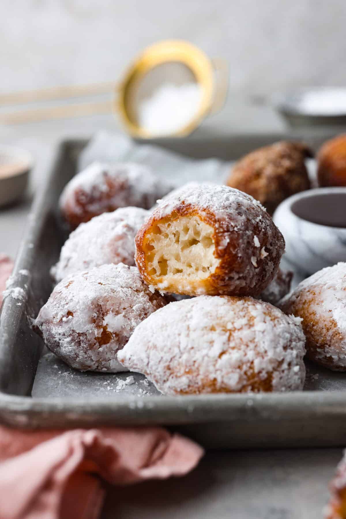 Side shot of zeppole coated in powdered sugar, one with a bite taken out of it. 