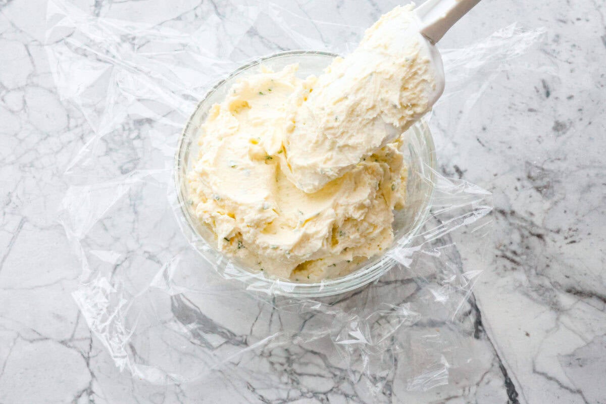 Overhead shot of spatula putting scoops of mixed Boursin cheese ingredients into a bowl lined with saran wrap. 