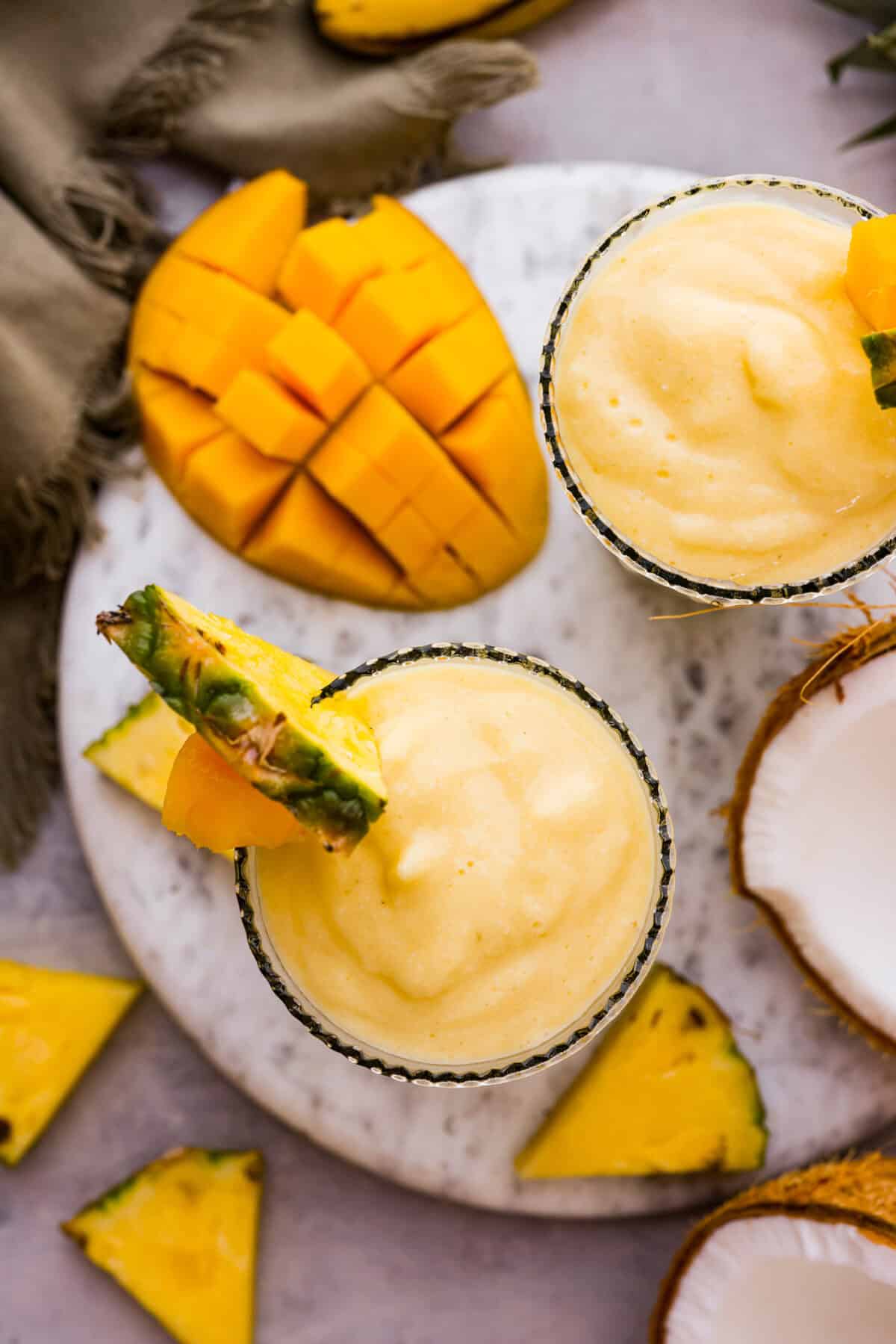 Overhead shot looking down at the top of two glasses next to pineapple wedges, cut up mango and half of a coconut. 