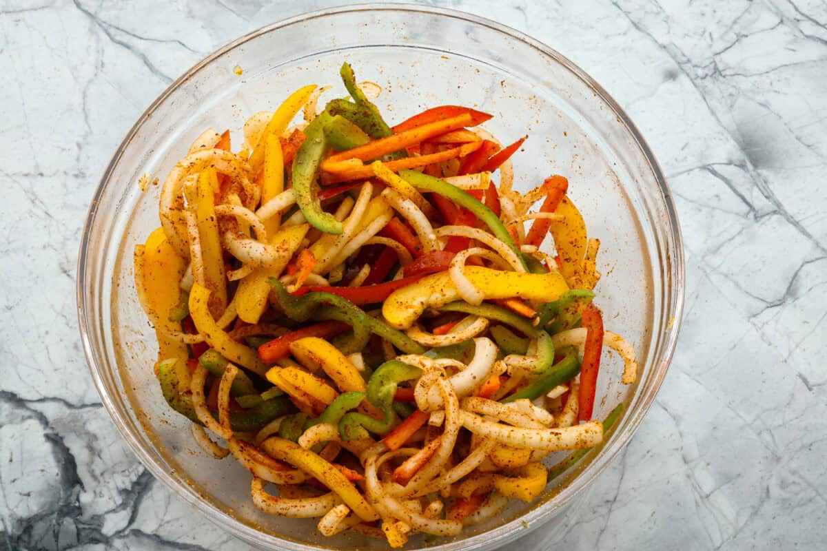 Overhead shot of sliced veggies coated in fajita seasonings in a glass bowl. 