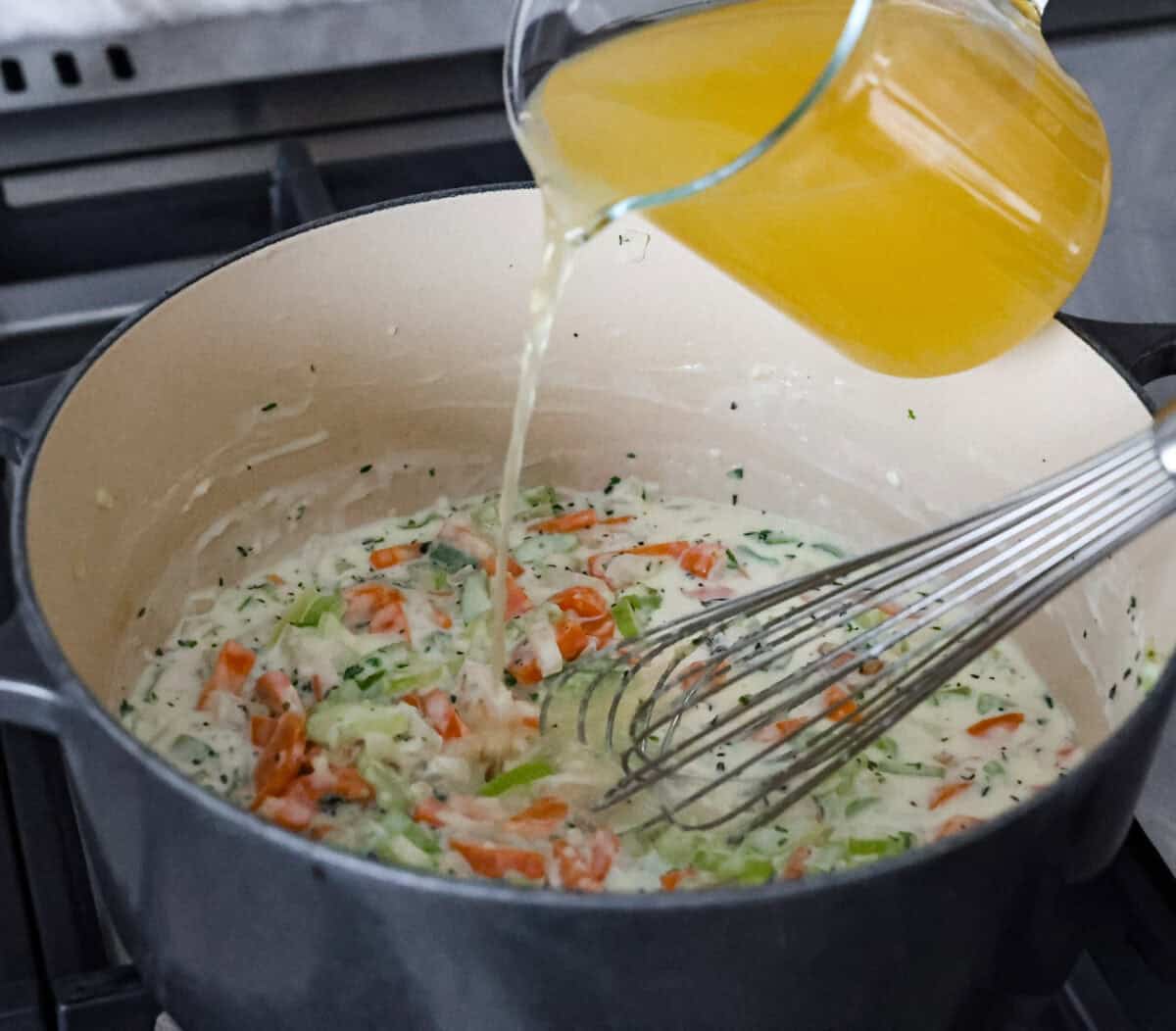 This creamy chicken noodle soup tastes exactly how it sounds! It's classic chicken noodle soup but creamy, rich and filling! Loaded with hearty chicken, tender carrots, supple noodles, and lots of delicious flavors, it's perfect for winter! Angle shot of someone pouring chicken broth into the vegetable and flour mixture.