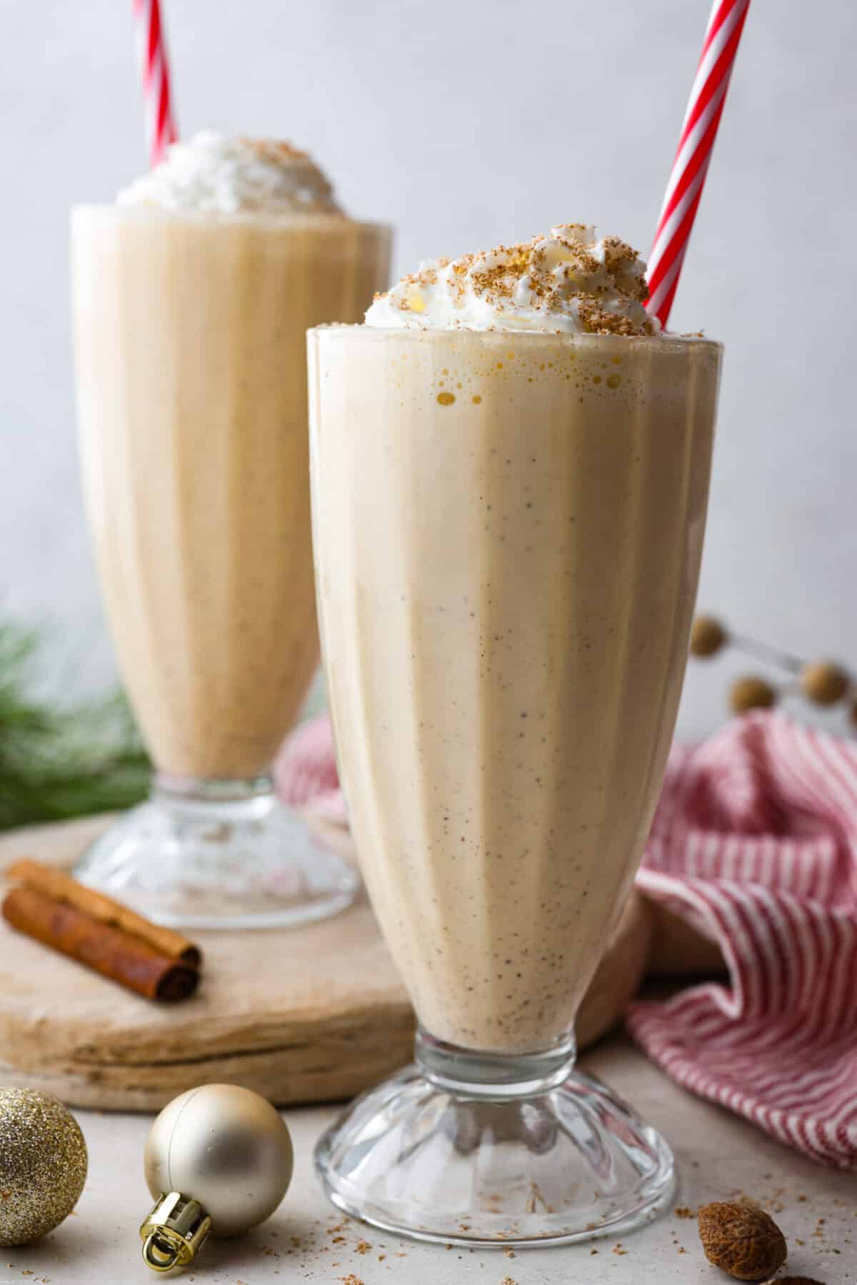 This creamy Eggnog Milkshake is decadent and super flavorful. It takes four simple ingredients and 5 minutes of your time. If you thought you liked eggnog before, you are going to absolutely love it after trying this shake. Side shot of two glasses full of eggnog milkshake with festive straw.
