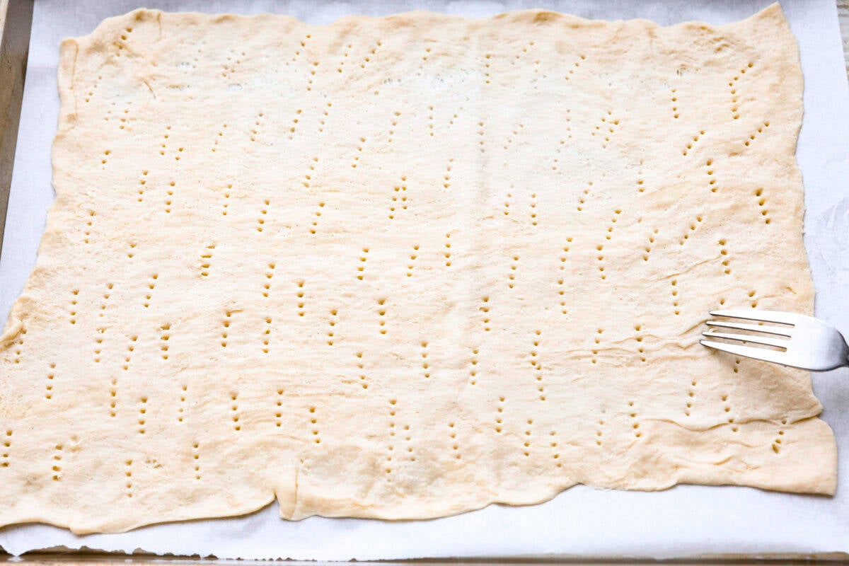 Maple Bacon Crack is as delicious and addicting as it sounds! It has flaky pastry dough drizzled in a sweet maple sauce and is loaded with crispy bacon pieces! It's the perfect sweet and salty snack! Overhead shot of crescent sheet dough being pricked a bunch with a fork.