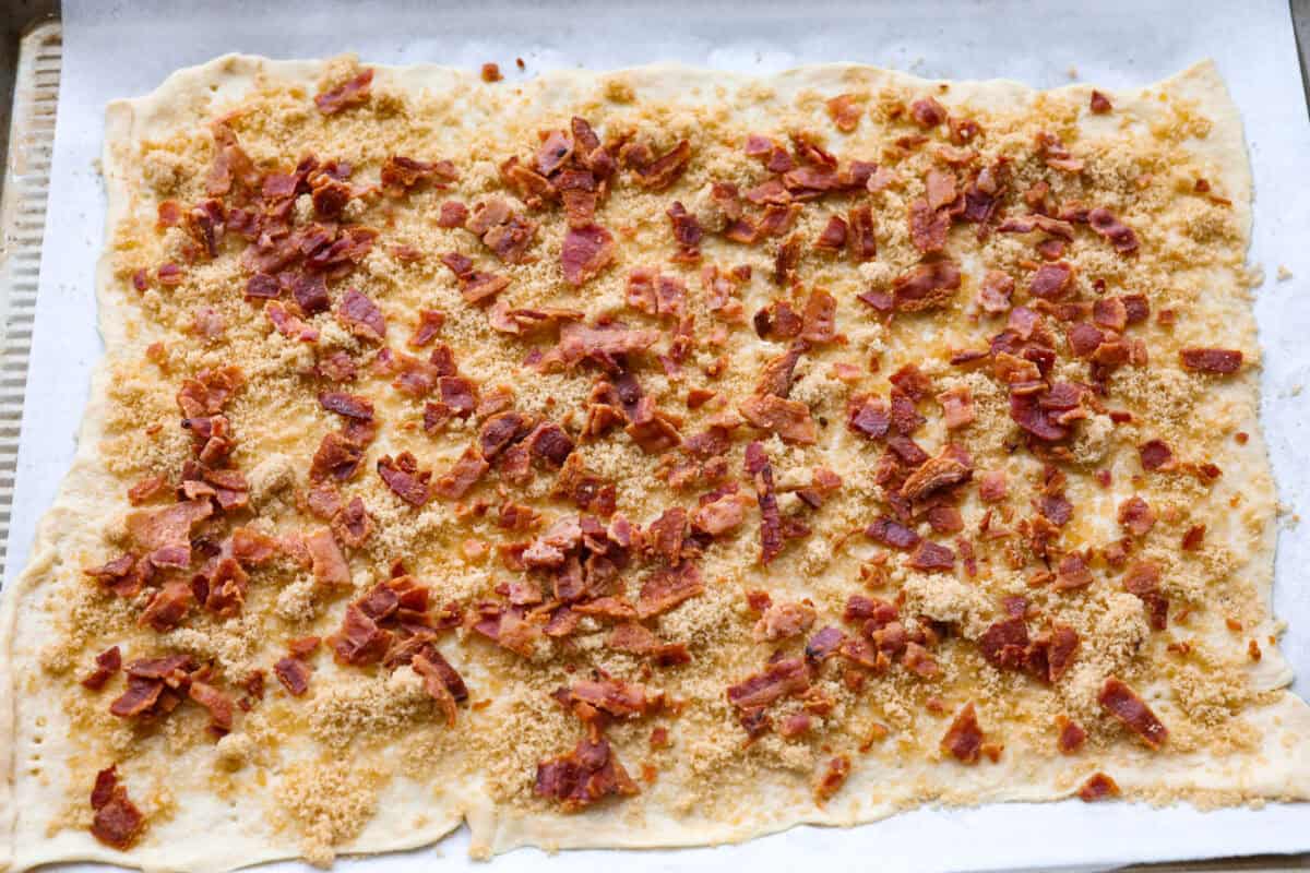 Maple Bacon Crack is as delicious and addicting as it sounds! It has flaky pastry dough drizzled in a sweet maple sauce and is loaded with crispy bacon pieces! It's the perfect sweet and salty snack! Overhead shot of bacon pieces over the brown sugar and maple on the crescent dough.