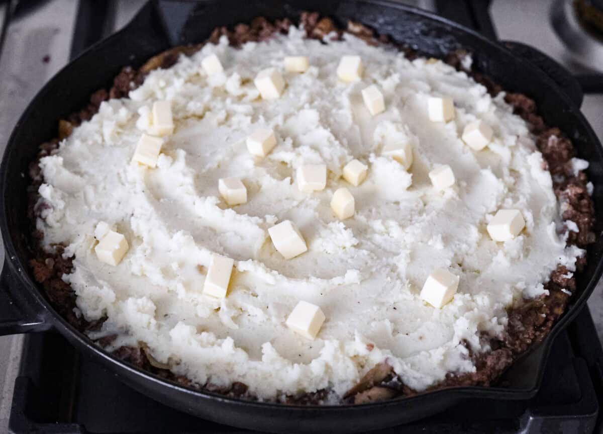 Angle shot of the mashed potatoes spread over the salisbury steak mixture with cubed butter on top. 